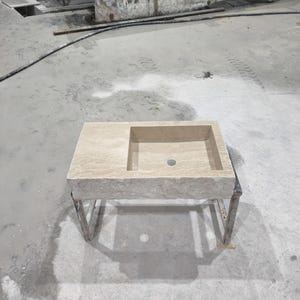 Könnte beinhalten: Ein beigefarbenes Steinklosterbecken mit einem rechteckigen Becken und einem einzigen Abflussloch. Das Waschbecken ist auf einem Metallgestell mit schlichtem Design montiert.