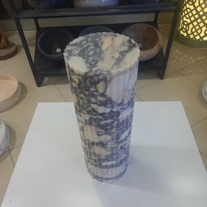 May include: A tall, cylindrical marble object with a fluted design. The stone features a mix of white, gray, and purple veining. The object is standing on a white surface, with other stone objects visible in the background.
