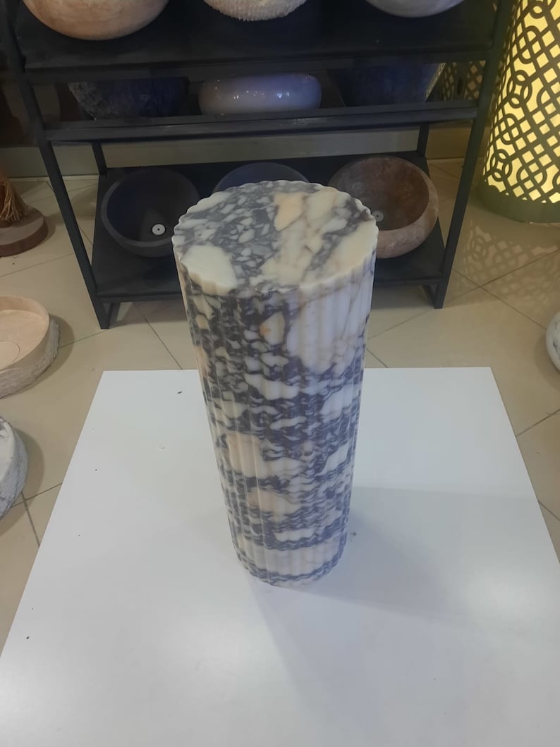 May include: A tall, cylindrical marble object with a fluted design. The stone features a mix of white, gray, and purple veining. The object is standing on a white surface, with other stone objects visible in the background.