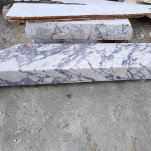 May include: A collection of marble slabs with a white base and striking purple veining. The slabs vary in size and shape, with a prominent rectangular piece in the foreground. The natural stone has a polished finish, showcasing its unique patterns.