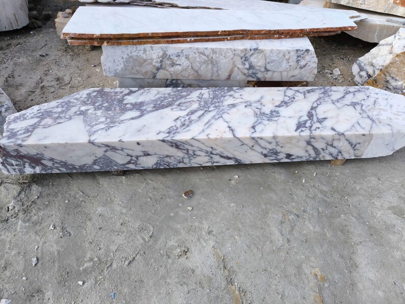 May include: A collection of marble slabs with a white base and striking purple veining. The slabs vary in size and shape, with a prominent rectangular piece in the foreground. The natural stone has a polished finish, showcasing its unique patterns.