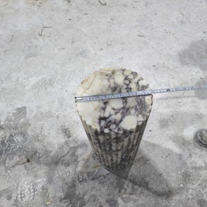 May include: A carved stone sculpture with a fluted, cone-like shape. The top is a circular shape with white and gray marbling. A measuring tape shows the diameter of the top. The sculpture is on a concrete surface.