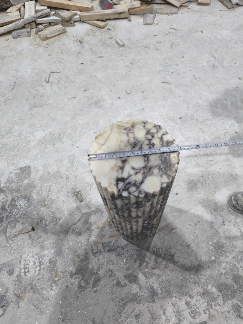 May include: A carved stone sculpture with a fluted, cone-like shape. The top is a circular shape with white and gray marbling. A measuring tape shows the diameter of the top. The sculpture is on a concrete surface.