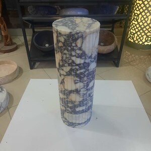 May include: A cylindrical marble pedestal with a fluted design. The stone features a mix of white, beige, and dark blue veining. The pedestal is standing on a white surface, with other stone bowls on a black metal shelf in the background.