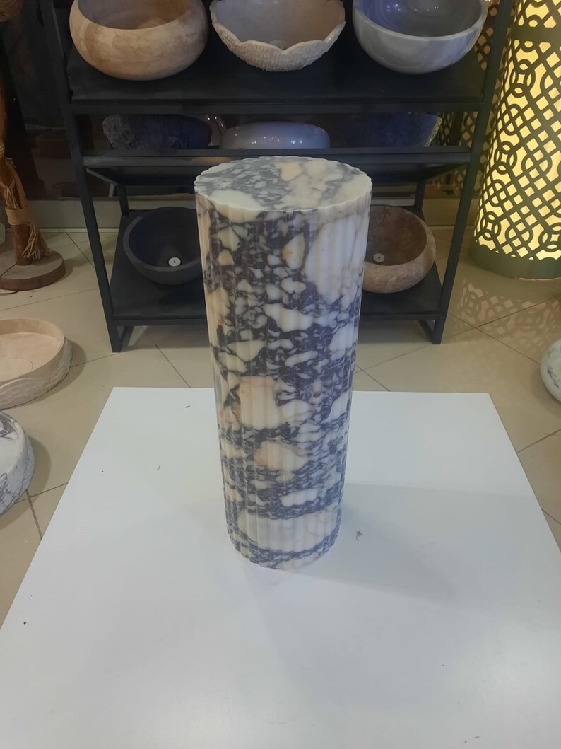 May include: A cylindrical marble pedestal with a fluted design. The stone features a mix of white, beige, and dark blue veining. The pedestal is standing on a white surface, with other stone bowls on a black metal shelf in the background.