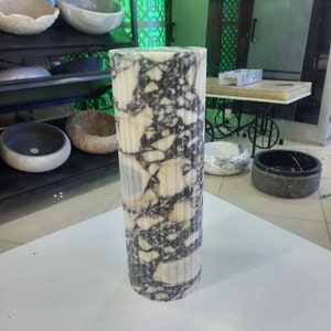May include: A tall, cylindrical marble pillar with vertical fluting. The pillar features a striking pattern of white, black, and tan veining. The stone column is a decorative element, likely for interior design or architectural use.