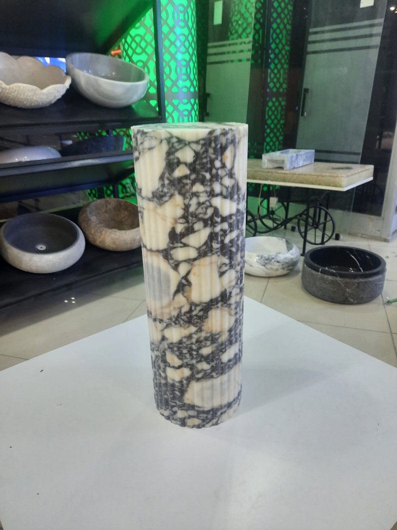 May include: A tall, cylindrical marble pillar with vertical fluting. The pillar features a striking pattern of white, black, and tan veining. The stone column is a decorative element, likely for interior design or architectural use.