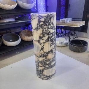 May include: A tall, cylindrical marble pillar with a fluted design. The pillar features a cream-colored base with dark gray and black veining throughout. The pillar is standing on a white surface, with other stone bowls visible in the background.