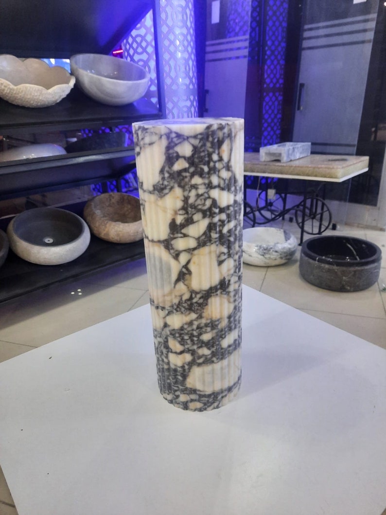 May include: A tall, cylindrical marble pillar with a fluted design. The pillar features a cream-colored base with dark gray and black veining throughout. The pillar is standing on a white surface, with other stone bowls visible in the background.