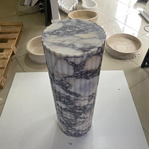 May include: A tall, cylindrical marble pedestal with a fluted design. The top is a circular shape with a wavy edge. The marble has a white base with gray and purple veining. The pedestal is on a white surface.
