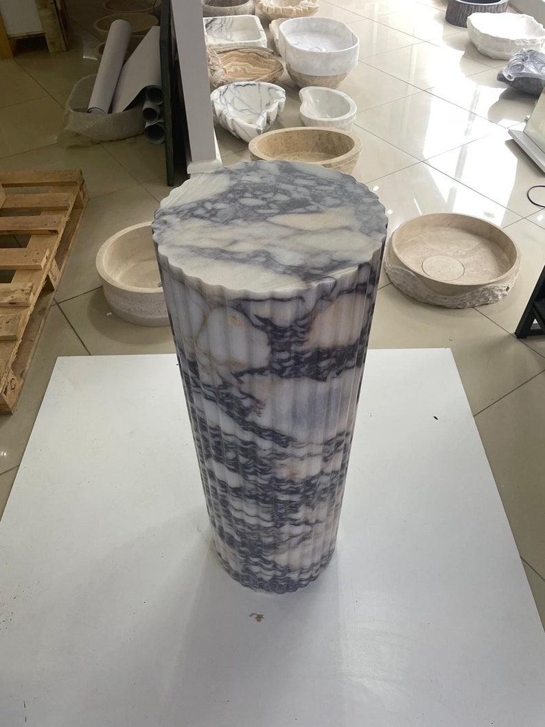 May include: A tall, cylindrical marble pedestal with a fluted design. The top is a circular shape with a wavy edge. The marble has a white base with gray and purple veining. The pedestal is on a white surface.