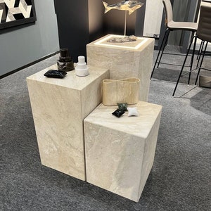 May include: Three square stone pedestals with a decorative fish sculpture on the top pedestal. The pedestals are made of a light beige stone with a marbled pattern. The fish sculpture is made of a dark gray material and has a silver finish.