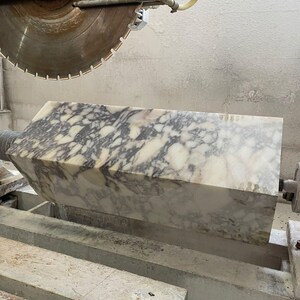 May include: A large, rectangular block of marble with a white and gray patterned surface is being cut by a circular saw. The saw blade is large and metallic, and the marble block is resting on a wooden support.