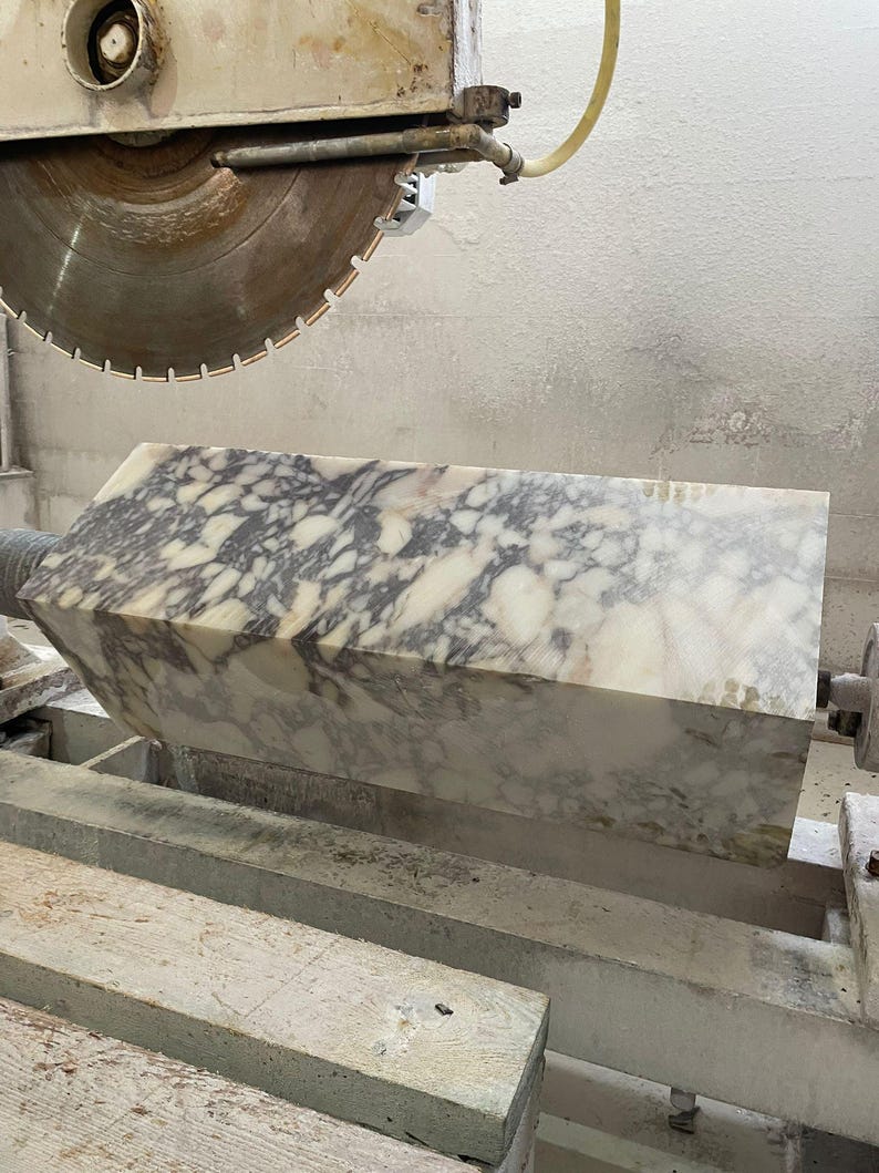 May include: A large, rectangular block of marble with a white and gray patterned surface is being cut by a circular saw. The saw blade is large and metallic, and the marble block is resting on a wooden support.