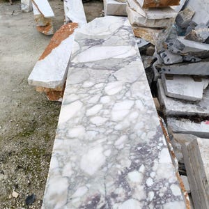 May include: A large, rectangular slab of marble with a complex pattern of gray and purple veining on a white background. The stone is surrounded by other marble blocks of various sizes and colors, including white, orange, and gray.