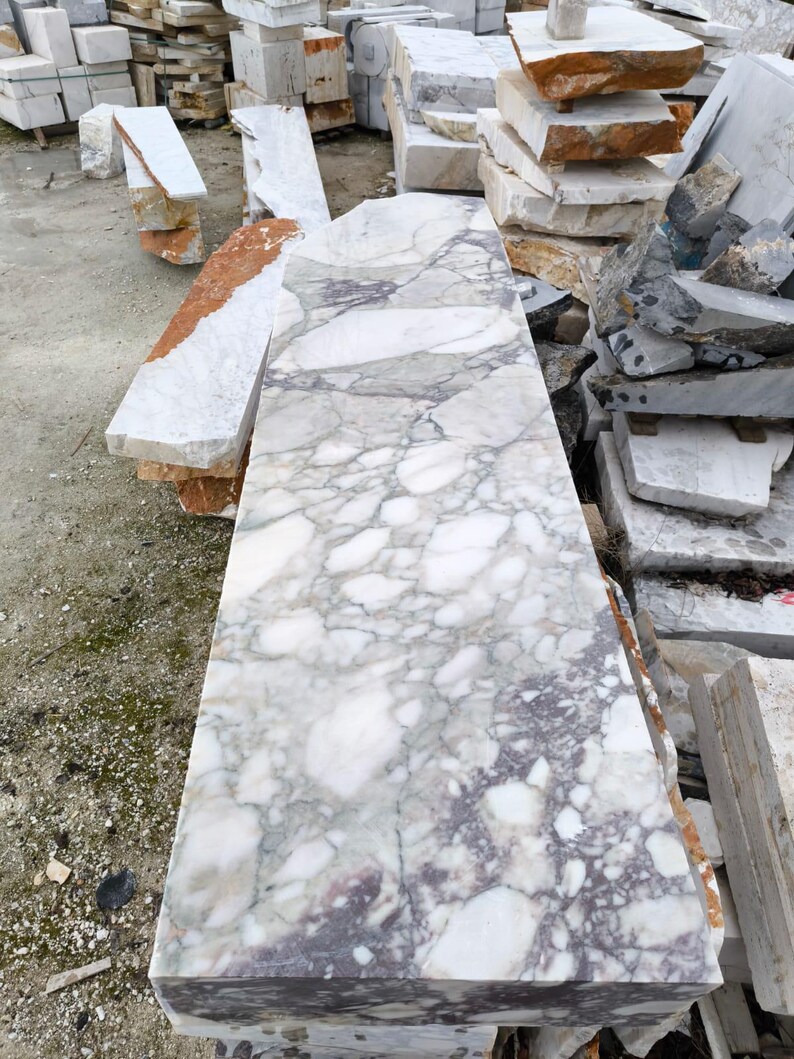 May include: A large, rectangular slab of marble with a complex pattern of gray and purple veining on a white background. The stone is surrounded by other marble blocks of various sizes and colors, including white, orange, and gray.