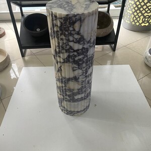 May include: A cylindrical marble pedestal with a fluted design. The pedestal is white with dark purple veining. It is standing on a white surface. Other stone bowls are visible in the background.