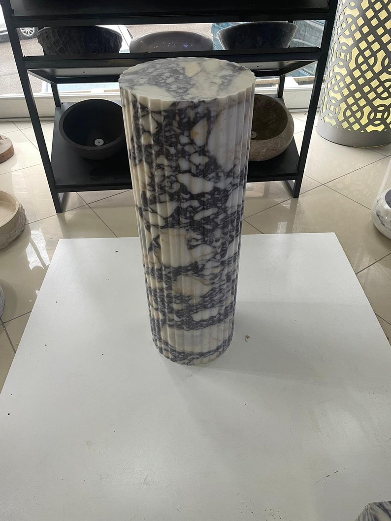 May include: A cylindrical marble pedestal with a fluted design. The pedestal is white with dark purple veining. It is standing on a white surface. Other stone bowls are visible in the background.