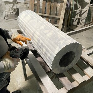 May include: A large, fluted marble column with a circular opening at the end. The column is white with gray veining and is being worked on with a power tool. The workshop setting includes wooden pallets and other stone elements.