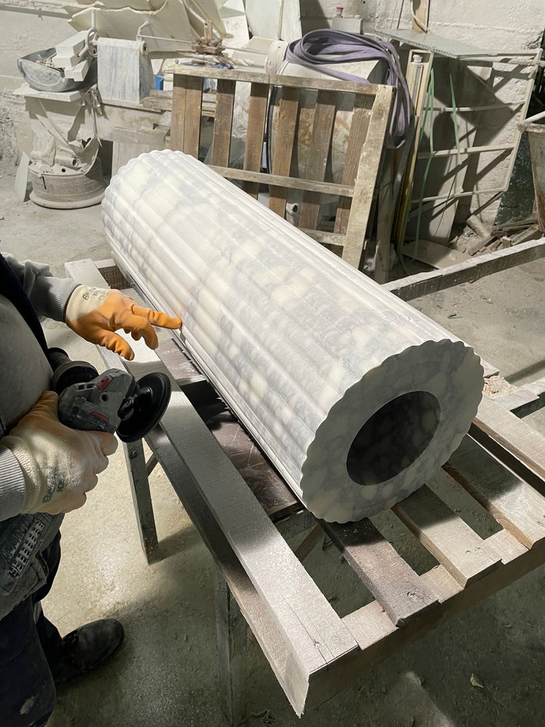 May include: A large, fluted marble column with a circular opening at the end. The column is white with gray veining and is being worked on with a power tool. The workshop setting includes wooden pallets and other stone elements.