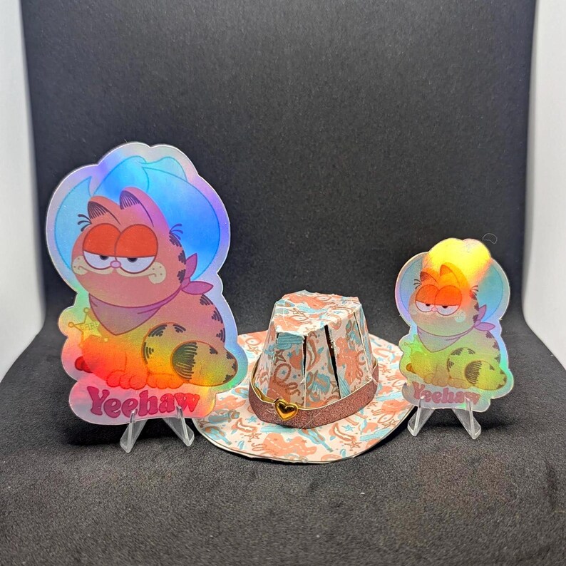 May include: Three holographic stickers featuring a cartoon cat wearing a cowboy hat and the text "Yeehaw". The stickers are on stands and in front of a small, colorful, cowboy hat.