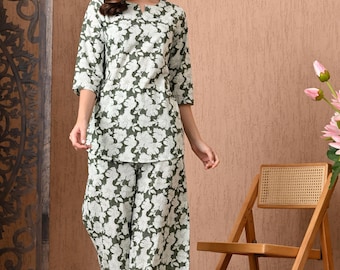 Può includere: Un completo a due pezzi con stampa floreale verde oliva e bianca. Il set comprende un top con maniche a tre quarti e pantaloni a gamba larga. La persona indossa il completo davanti a una sedia di legno e un paravento decorativo.