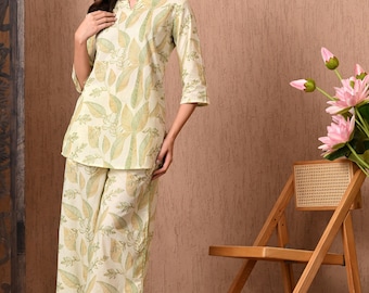 Puede incluir: Conjunto de pijama de dos piezas en color crema con un estampado de hojas verdes. La parte superior tiene un escote en pico y mangas tres cuartos. Los pantalones a juego son de pernera ancha. Una silla de madera y flores rosas están en el fondo.