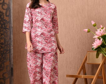 Puede incluir: Conjunto de pijama rosa con estampado floral. El conjunto incluye una parte superior con mangas tres cuartos y pantalones de pernera ancha. La modelo lleva el conjunto delante de una silla de madera y una pantalla decorativa.