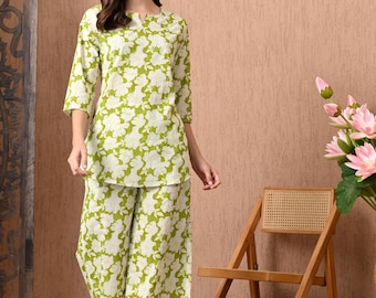 Puede incluir: Un conjunto de dos piezas con estampado floral en verde y blanco. El conjunto incluye una túnica con mangas tres cuartos y pantalones de pernera ancha. El atuendo se usa con sandalias. Una silla de madera y flores rosas están en el fondo.
