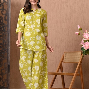 May include: A two-piece floral print outfit in a light green hue. The set includes a three-quarter sleeve top and wide-leg pants, both adorned with white floral patterns. The outfit is worn by a person. A wooden chair and pink flowers are in the background.