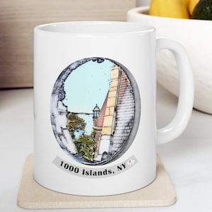 May include: White ceramic mug featuring a circular image of a scenic view with a tower and water. The image is framed by a decorative border. The text "1000 Islands, NY" is printed below the image. The mug has a curved handle.