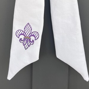 May include: White fabric door wreath bow with a purple fleur-de-lis design.