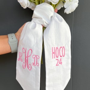 May include: White bouquet of flowers tied with a white satin ribbon. The ribbon is embroidered with the initials "K.H.D." and the text "HOCO 24".