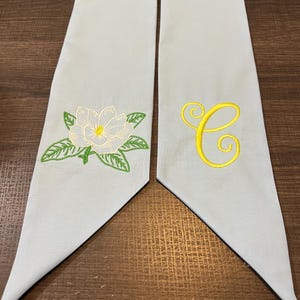 May include: Two light blue graduation stoles. One stole features a white magnolia flower with green leaves, and the other has a yellow embroidered letter "C". The stoles are made of a soft fabric.