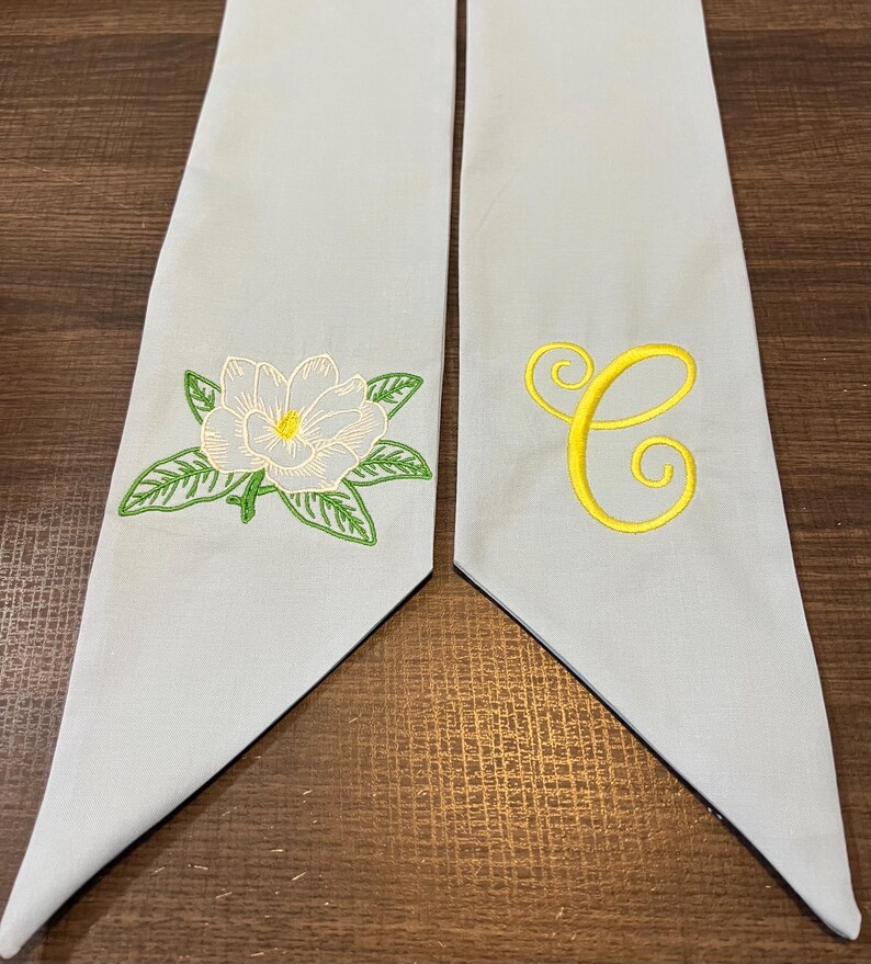 May include: Two light blue graduation stoles. One stole features a white magnolia flower with green leaves, and the other has a yellow embroidered letter "C". The stoles are made of a soft fabric.
