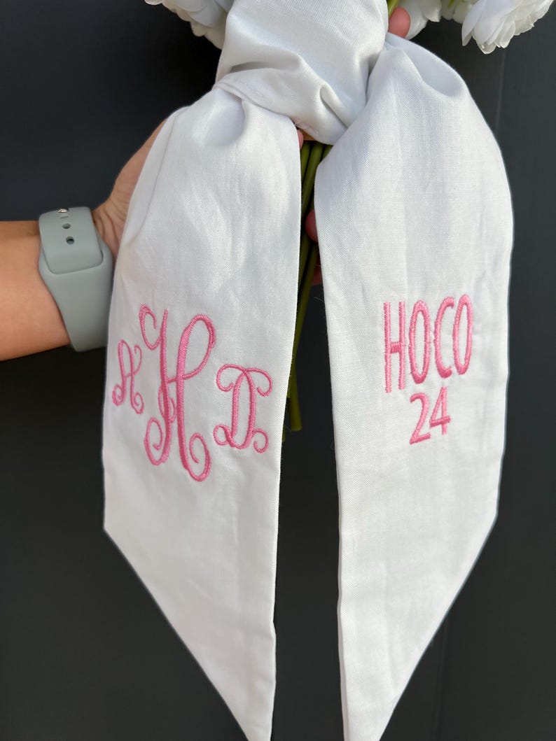 May include: White sash with pink embroidered text. One side features the initials "HD" in a cursive font. The other side reads "HOCO 24" in a block font. The sash is held in front of a dark background.