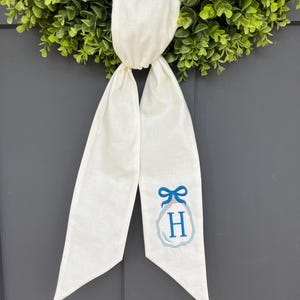 May include: A white fabric wreath sash with a blue embroidered initial "H" and bow design. The sash is hanging in front of a green wreath. The background is a gray door.
