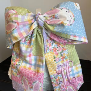 K&ouml;nnte beinhalten: Eine gro&szlig;e, farbenfrohe Stoffschleife mit einem Patchwork-Design in Pastellfarben. Die Schleife hat eine Hasenapplikation und einen gestickten Buchstaben "M". Die Schleife ist in der Mitte gebunden und dient wahrscheinlich zur Dekoration.