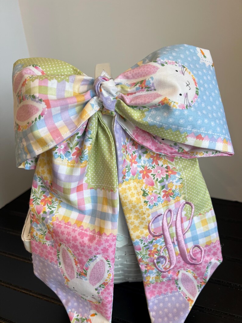 K&ouml;nnte beinhalten: Eine gro&szlig;e, farbenfrohe Stoffschleife mit einem Patchwork-Design in Pastellfarben. Die Schleife hat eine Hasenapplikation und einen gestickten Buchstaben "M". Die Schleife ist in der Mitte gebunden und dient wahrscheinlich zur Dekoration.