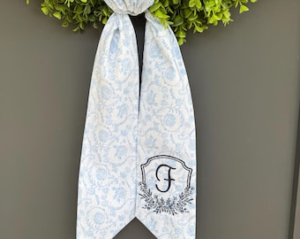 May include: A light blue and white floral patterned ribbon with the letter "F" embroidered on a decorative emblem. The ribbon is attached to a green wreath, creating a decorative accent for a door or wall.