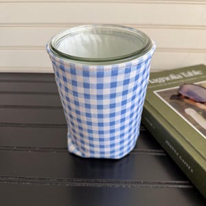 May include: A clear glass jar encased in a fabric sleeve featuring a blue and white gingham check pattern. The sleeve fits snugly around the exterior of the cylindrical glass container, which has a smooth, open top rim and a flat base.