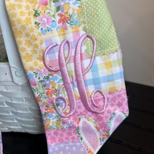K&ouml;nnte beinhalten: Ein farbenfrohes Stoffst&uuml;ck zum Thema Ostern mit Patchwork-Design. Der Stoff weist Blumen-, Tupfen- und Vichy-Muster in Pastellfarben auf. Ein rosa gestickter Buchstabe "M" ist deutlich zu sehen, mit Hasenohren am unteren Rand.