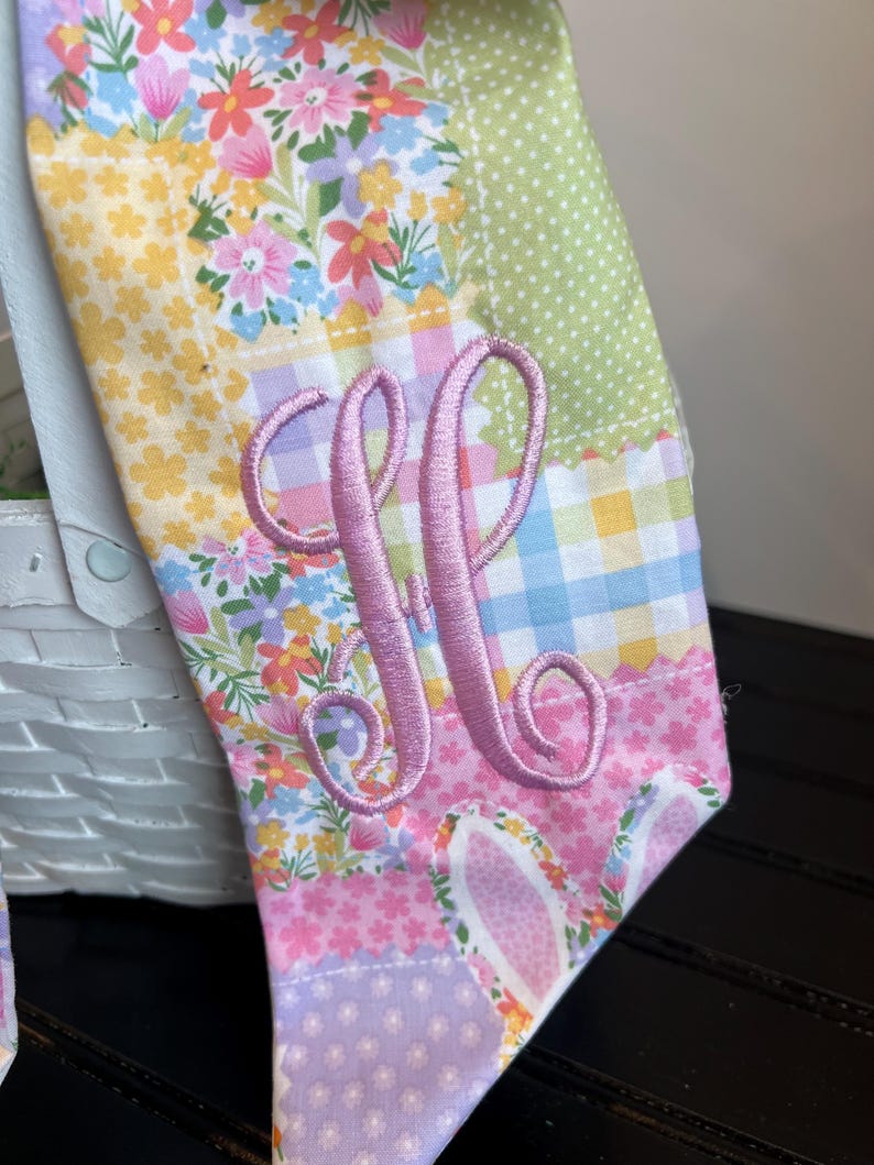 K&ouml;nnte beinhalten: Ein farbenfrohes Stoffst&uuml;ck zum Thema Ostern mit Patchwork-Design. Der Stoff weist Blumen-, Tupfen- und Vichy-Muster in Pastellfarben auf. Ein rosa gestickter Buchstabe "M" ist deutlich zu sehen, mit Hasenohren am unteren Rand.