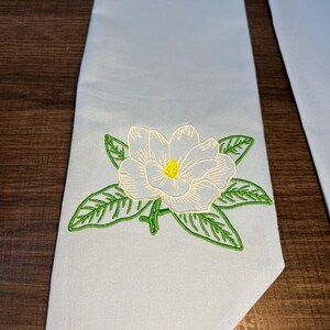 May include: Light blue graduation sash with an embroidered white magnolia flower with a yellow center and green leaves. The sash is made of a smooth fabric and is likely used for academic ceremonies.