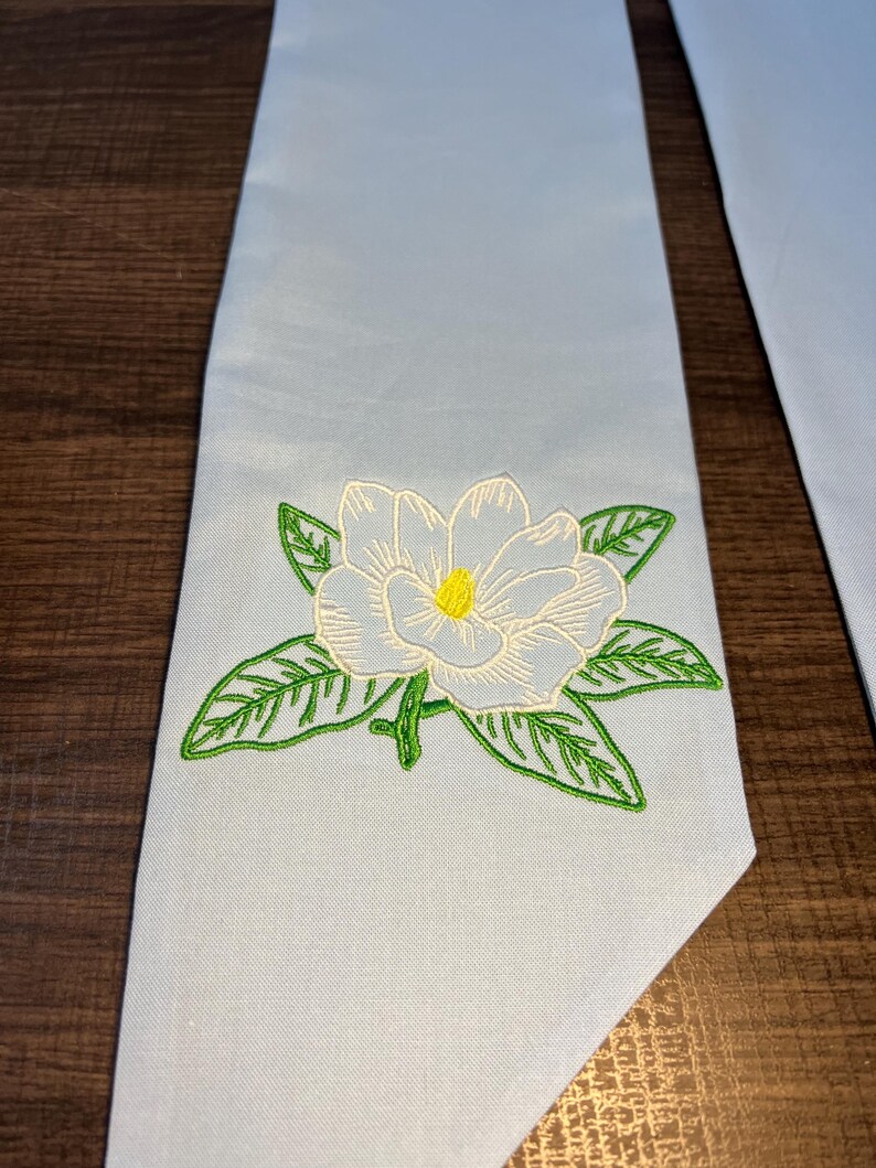 May include: Light blue graduation sash with an embroidered white magnolia flower with a yellow center and green leaves. The sash is made of a smooth fabric and is likely used for academic ceremonies.
