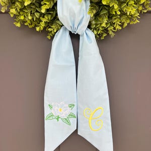 May include: A light blue wreath sash with a magnolia flower and the letter "C" embroidered in yellow. The sash is tied to a green wreath, creating a decorative accent for a door or wall.