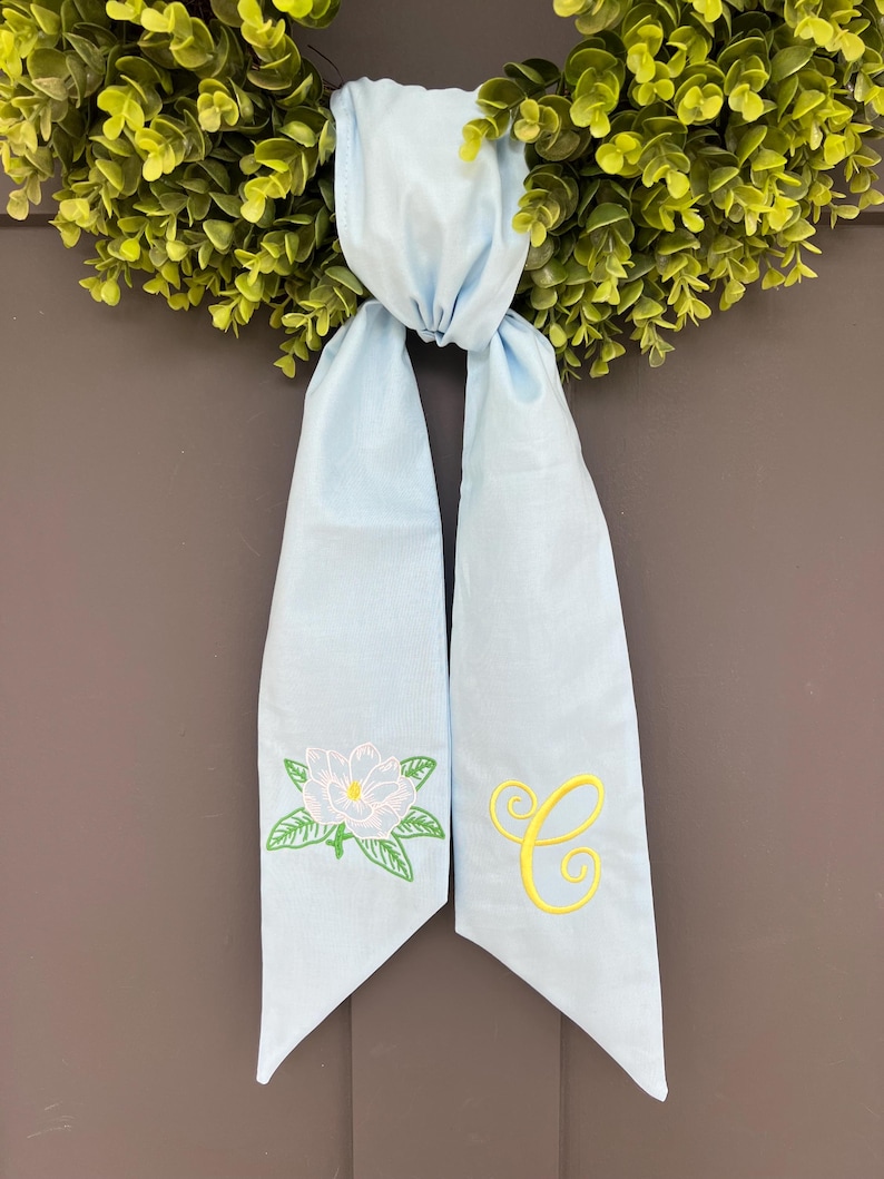 May include: A light blue wreath sash with a magnolia flower and the letter "C" embroidered in yellow. The sash is tied to a green wreath, creating a decorative accent for a door or wall.