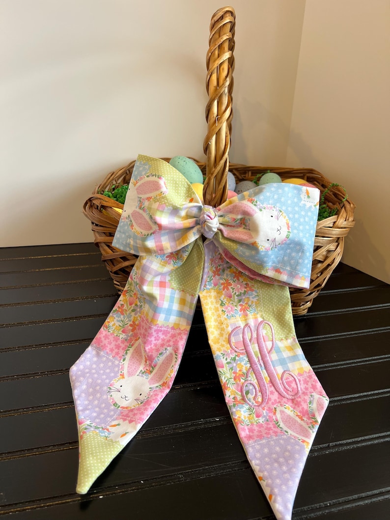 K&ouml;nnte beinhalten: Ein geflochtener Osterkorb aus Weide mit einer dekorativen Stoffschleife. Die Schleife hat ein Patchwork-Design mit Hasen- und Blumenmustern in Pastellfarben. Der Korb ist mit dekorativen Eiern gef&uuml;llt.