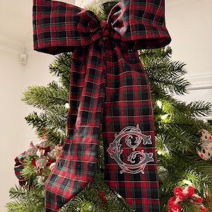 May include: A large, decorative Christmas tree bow in a classic red, black, and green plaid pattern. The bow is placed on a Christmas tree and features a white embroidered initial "C". The bow's long tails cascade down the tree.