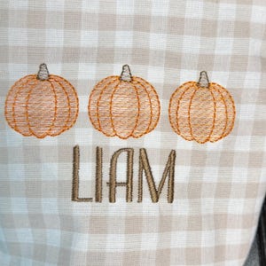 May include: A beige and white checkered fabric with three embroidered orange pumpkins and the name "LIAM" in brown thread. The pumpkins are detailed with stitched lines and stems.